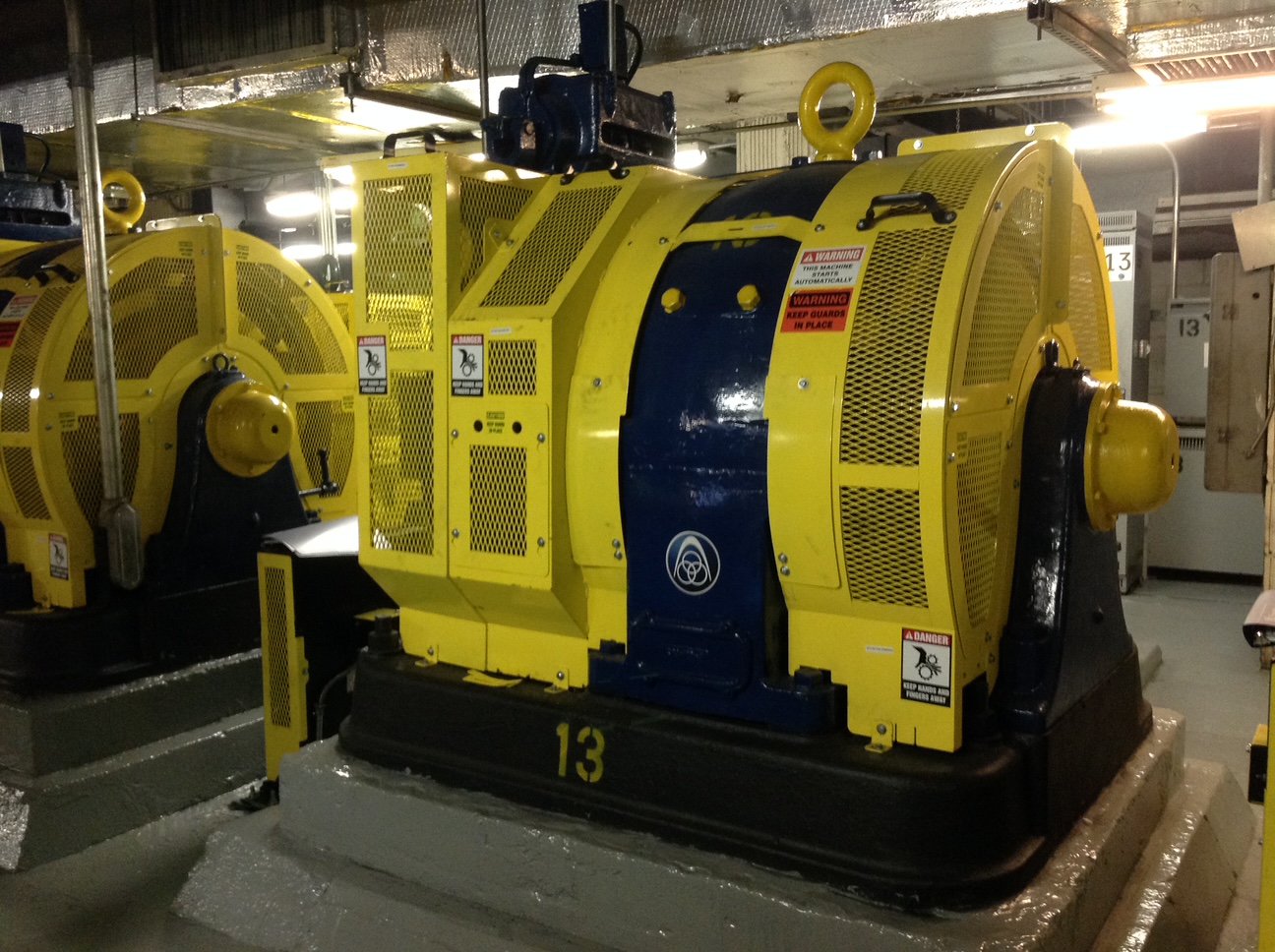 Yellow barrier guarding around an elevator traction machine and its sheave in a machine room, with keep-guards-in-place warning labels.
