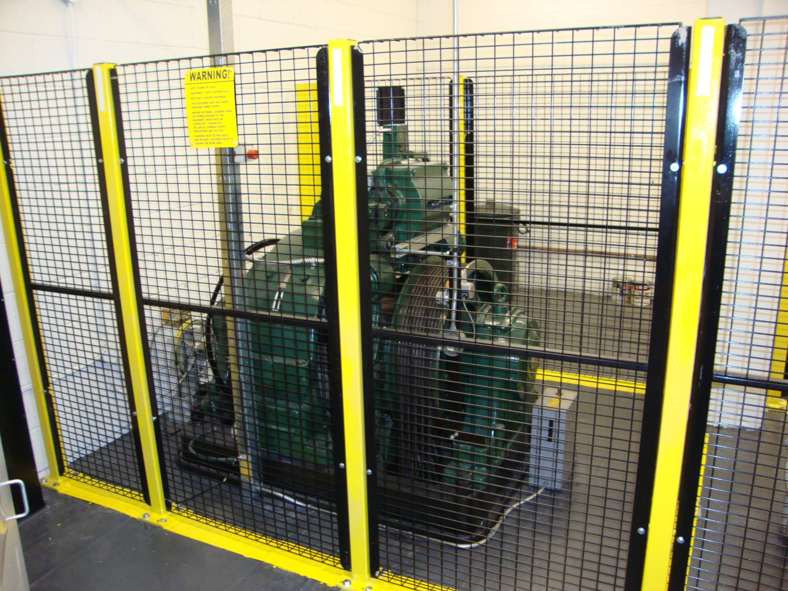 Welded wire-mesh perimeter guarding with yellow posts enclosing rotating machinery against a block wall.