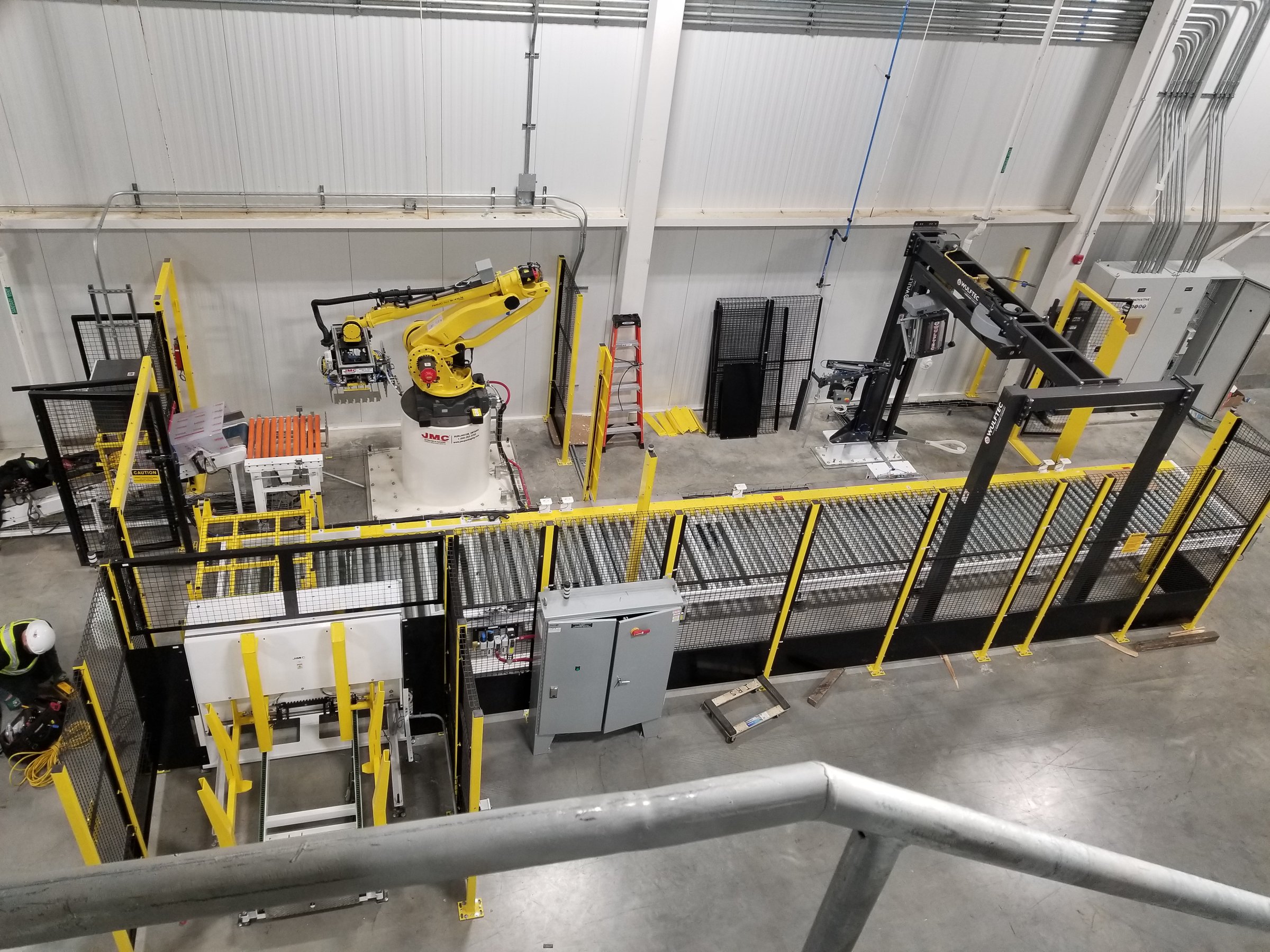 Overhead view of a robotic packaging cell enclosed by yellow wire mesh perimeter guarding, with a roller conveyor and packaging equipment beyond the barrier